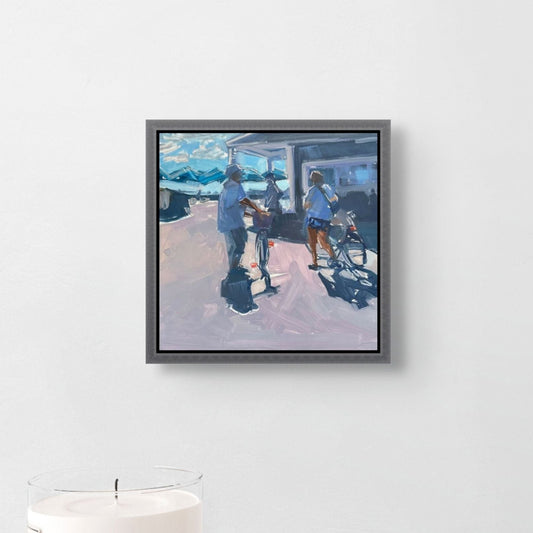 Framed painting of people with bicycles on a white wall above a white table with a candle.