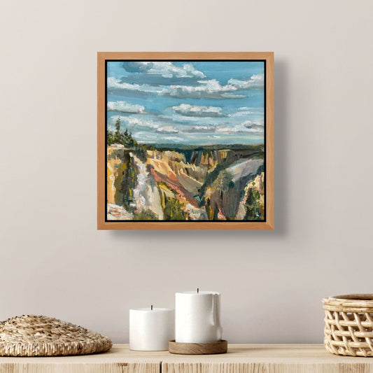 Framed landscape painting of a canyon on a wall above a wooden shelf with candles and baskets.