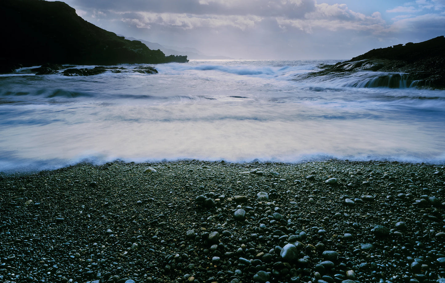 Stony beach with waves crashing onto the shore under a cloudy sky.