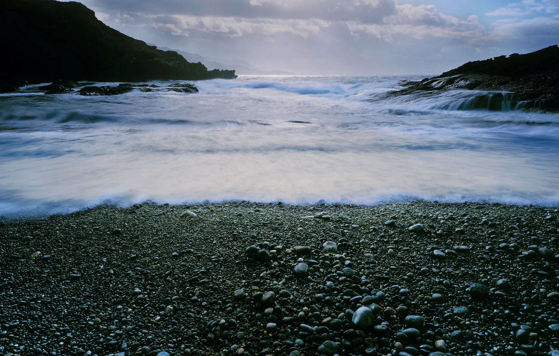 Stony beach with waves crashing onto the shore under a cloudy sky.