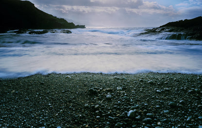 Stony beach with waves crashing onto the shore under a cloudy sky.