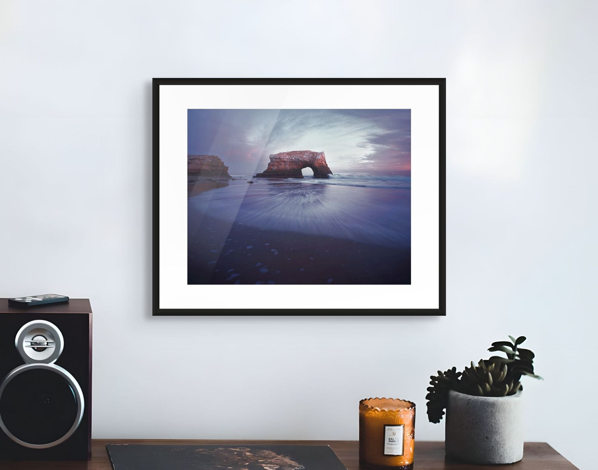 Framed artwork of a coastal scene with a rock formation on a wall above a table with decor items.